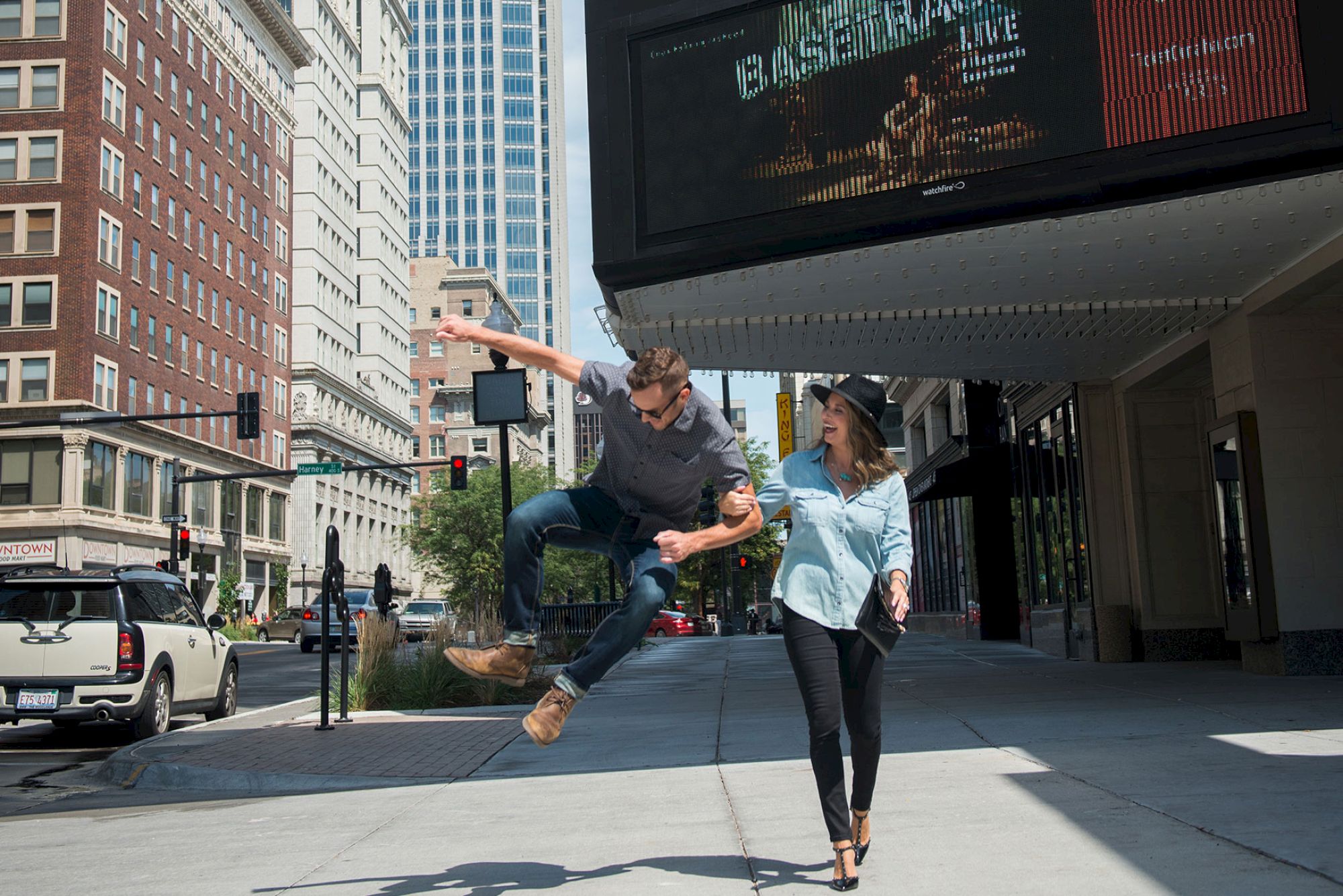 A man playfully jumps in the air alongside a woman walking on a city sidewalk near a theater marquee.