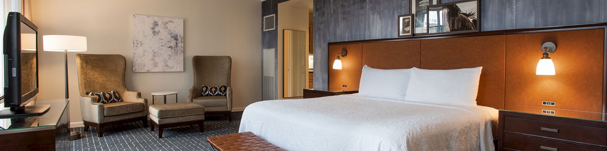 A modern hotel room with a king-size bed, upholstered chairs, a TV, and stylish decor, featuring a blue and brown color scheme.