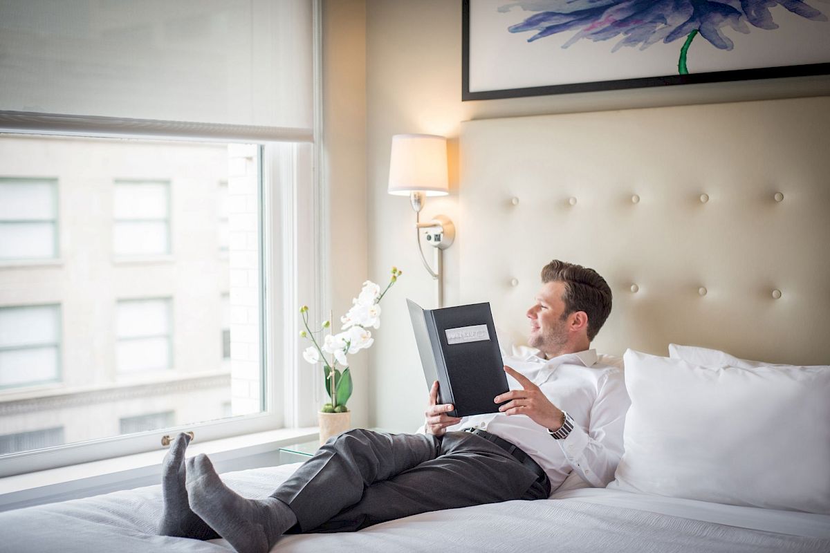 A person in a white shirt is lying on a bed, reading a book near a window with city views and a vase of flowers beside them.