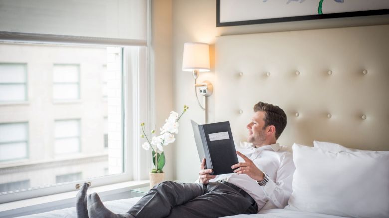 A person in a white shirt is lying on a bed, reading a book near a window with city views and a vase of flowers beside them.
