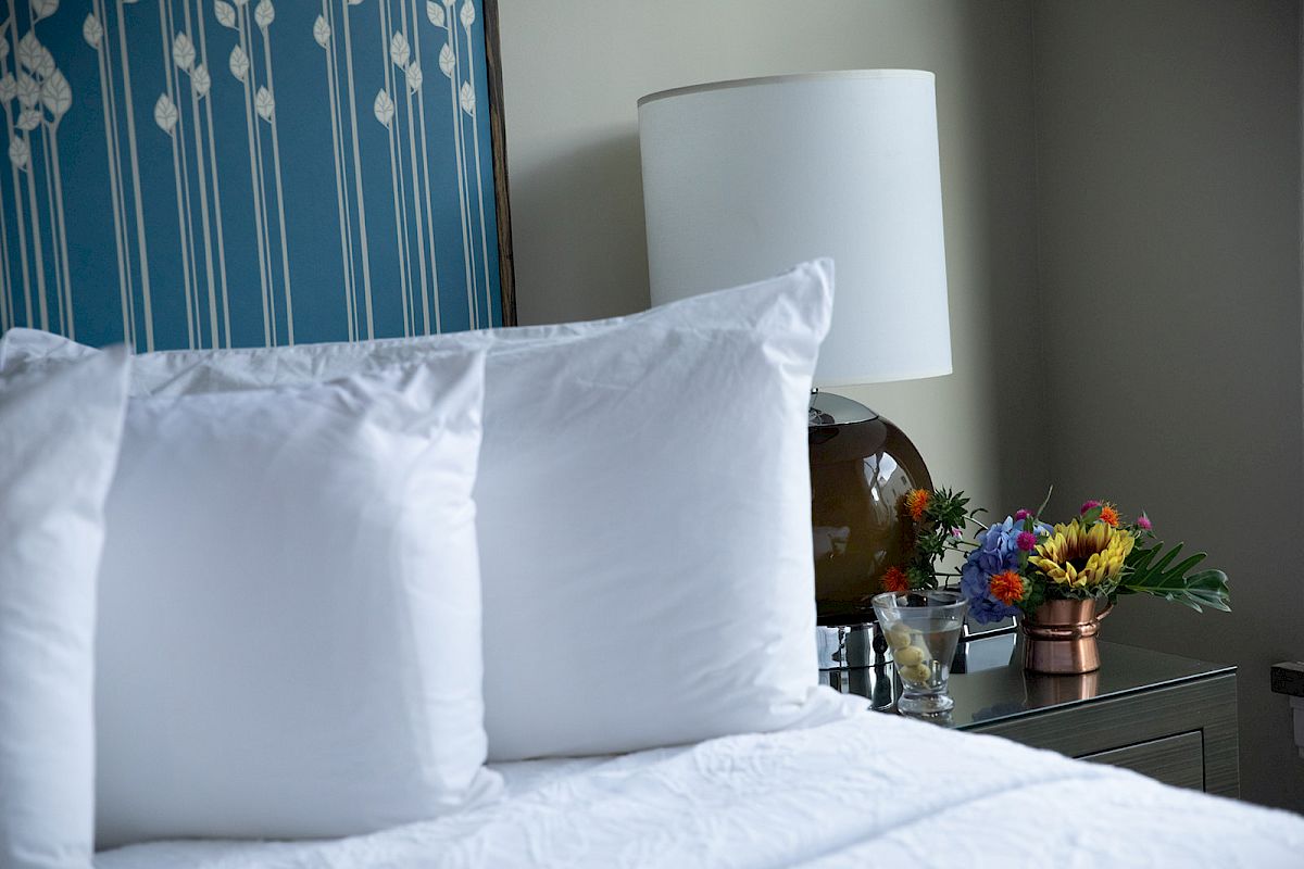 A neatly made bed with white pillows, a bedside table with a lamp, and a colorful floral arrangement in a small pot.