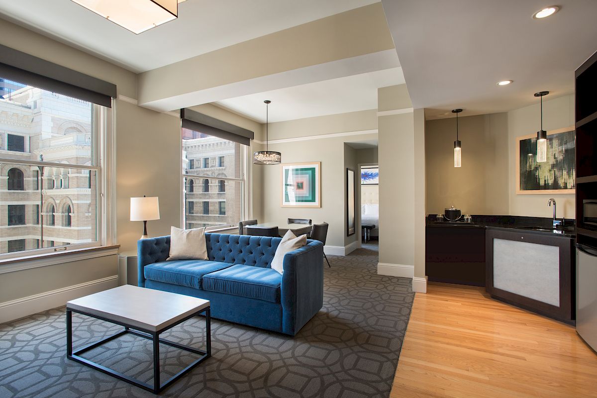 A modern living room and kitchen area with a blue sofa, table, art, and large windows with city views.