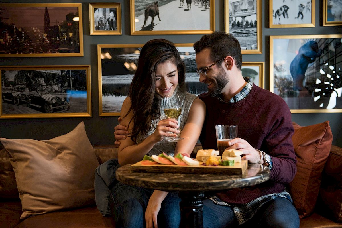 A couple sits together at a cozy café, smiling and enjoying drinks and a platter of snacks, surrounded by framed pictures on the wall.