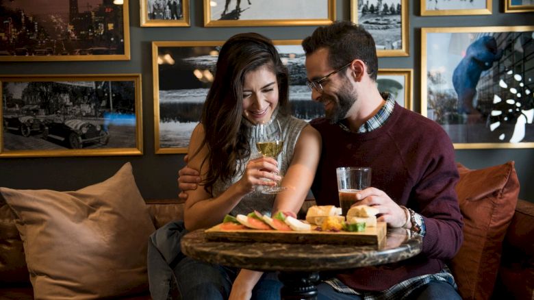 A couple sits together at a cozy café, smiling and enjoying drinks and a platter of snacks, surrounded by framed pictures on the wall.