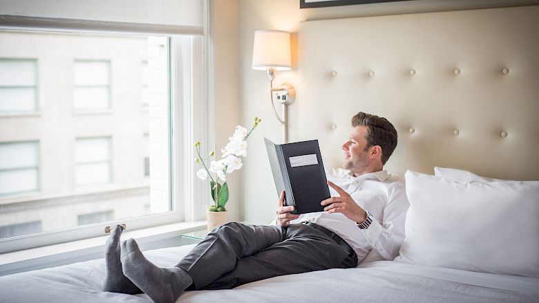 A person is relaxing on a bed, reading a book near a window with flowers in the background.