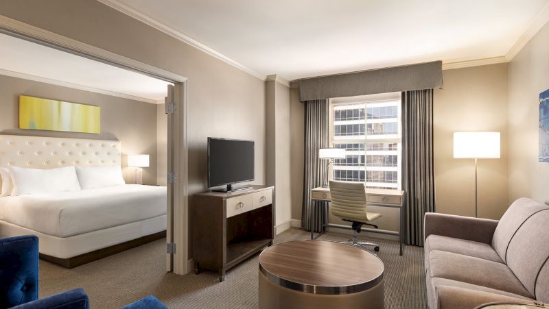 A hotel suite with a king-sized bed, sofa, desk, TV, and window