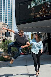A man joyfully jumps in the air on a city sidewalk, while a smiling woman walks beside him, both in casual attire.