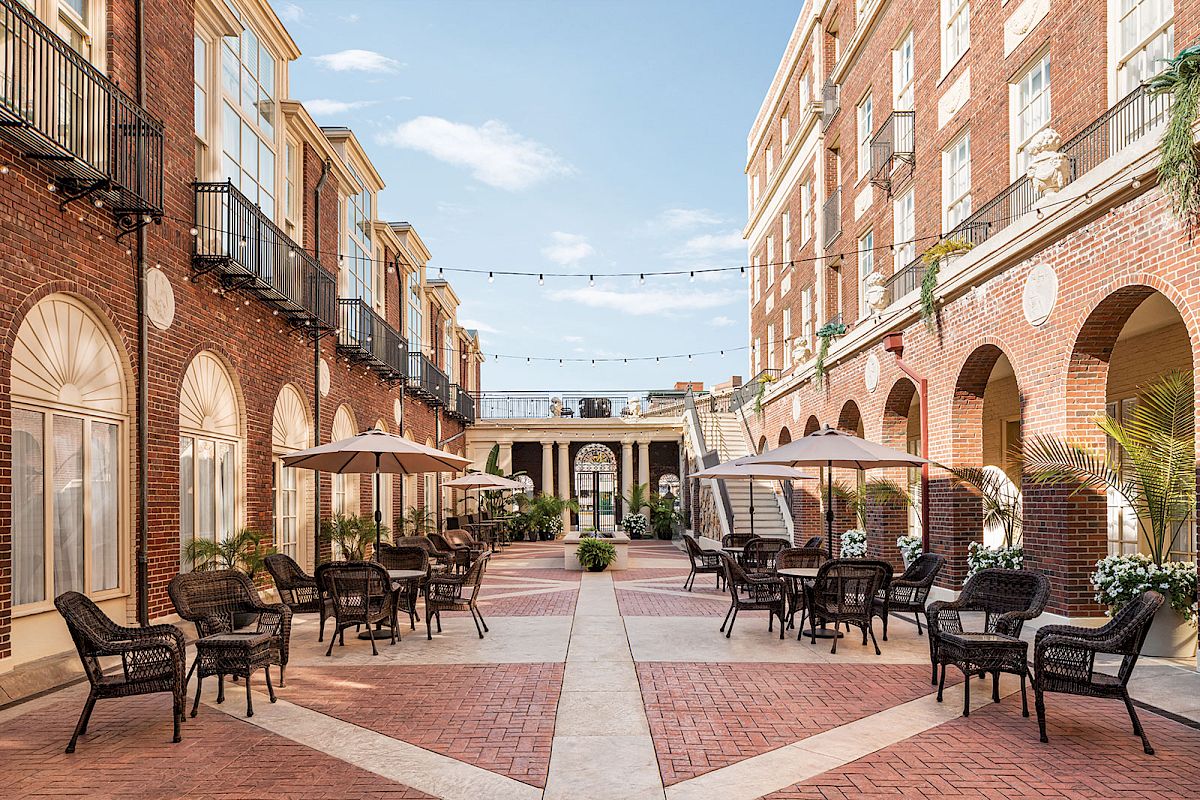 The image shows an elegant courtyard with brick buildings, outdoor seating, umbrellas, and string lights under a clear blue sky.