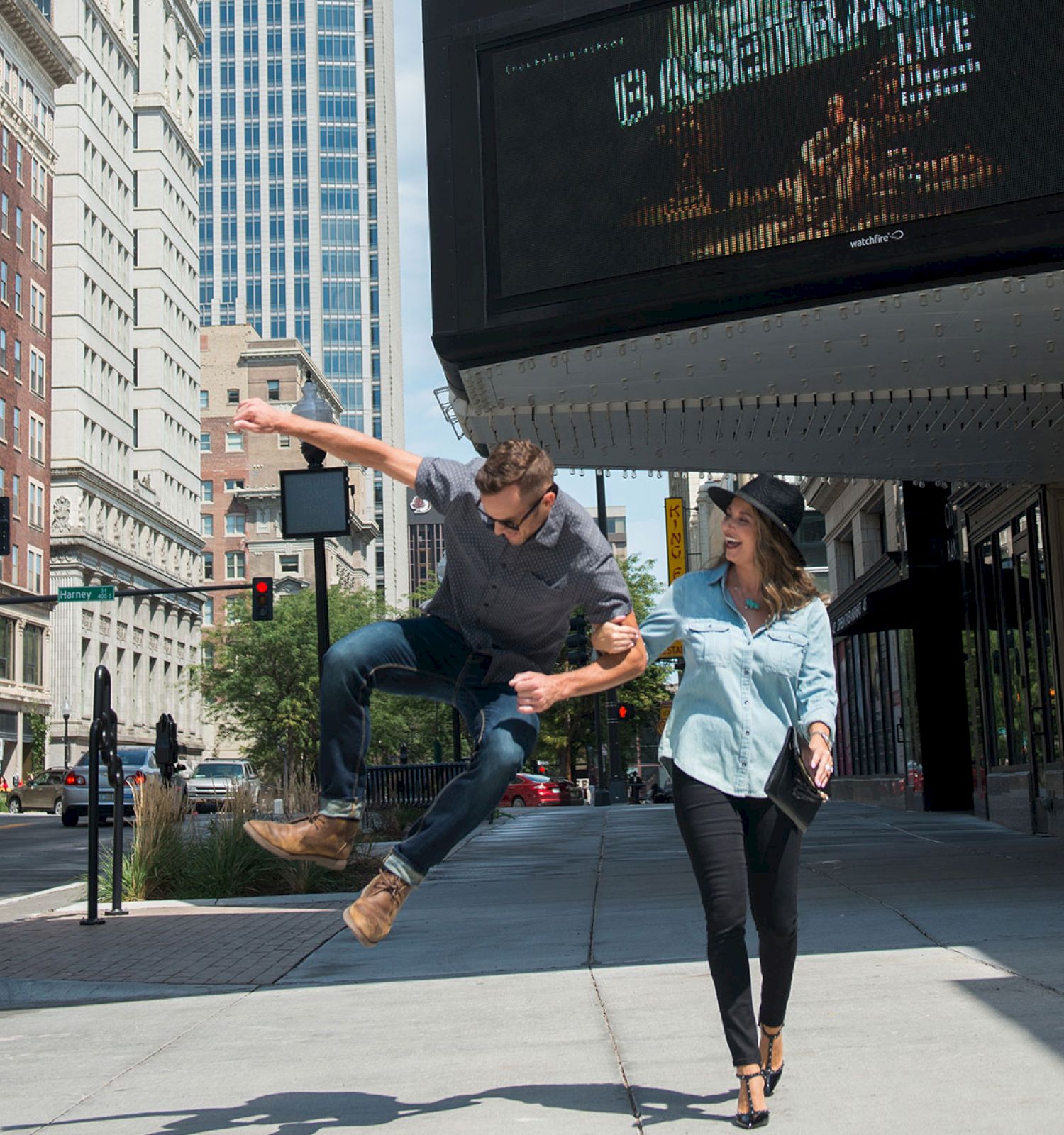 A man is jumping playfully while holding hands with a woman on a city sidewalk, with buildings and a theater marquee in the background.