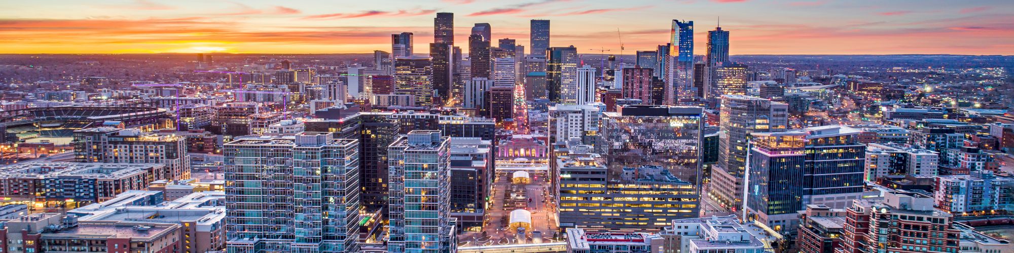 A city skyline at sunset with colorful skies, featuring numerous buildings, skyscrapers, and a park in the foreground.