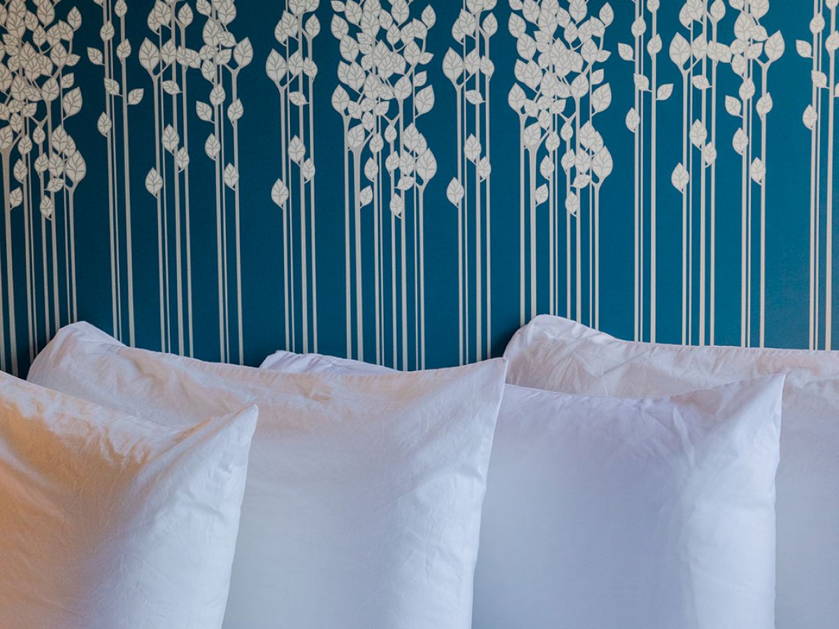 A bed with white pillows against a teal wall decorated with a pattern of white flowers on tall stems.