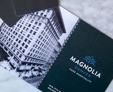 Promotional material for Magnolia Denver, featuring a historic building image and brand logo with contact details on a marble surface.