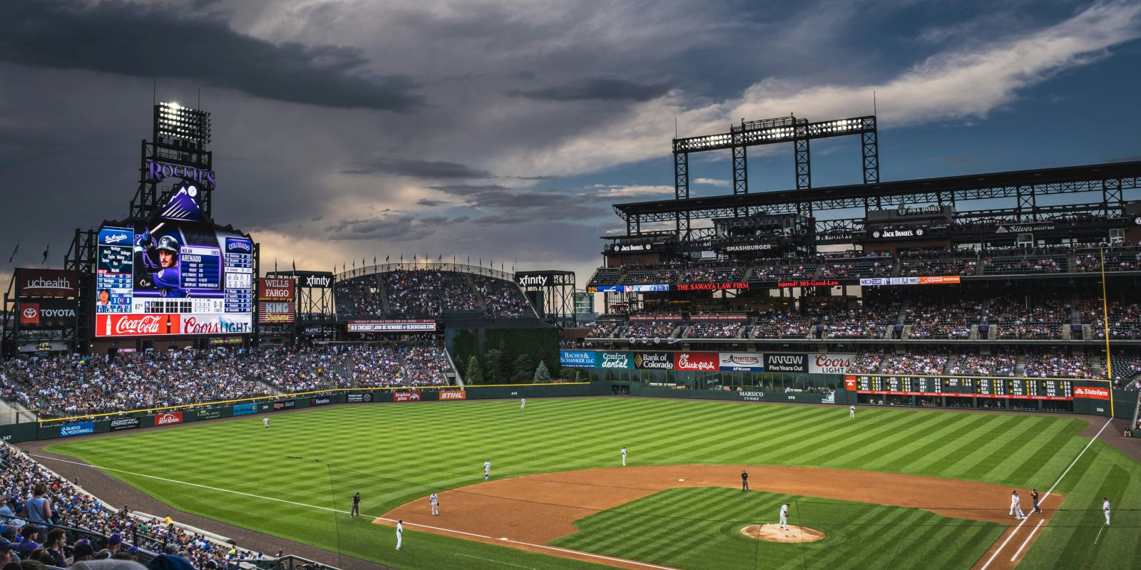 A baseball game at a large stadium with players on the field, an electronic scoreboard, and a crowd in the stands under a cloudy sky.
