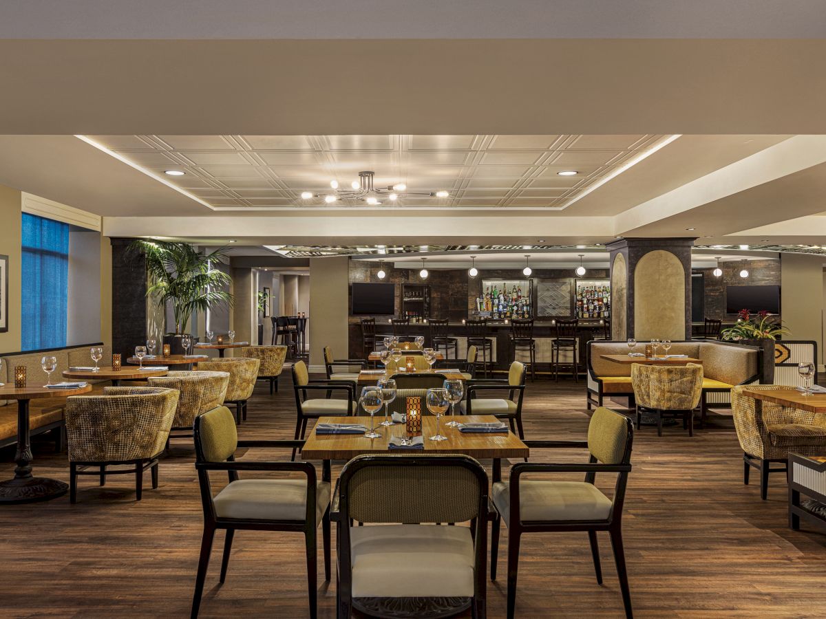The image shows an upscale dining area with elegant tables and chairs, softly lit ambiance, and a bar in the background.