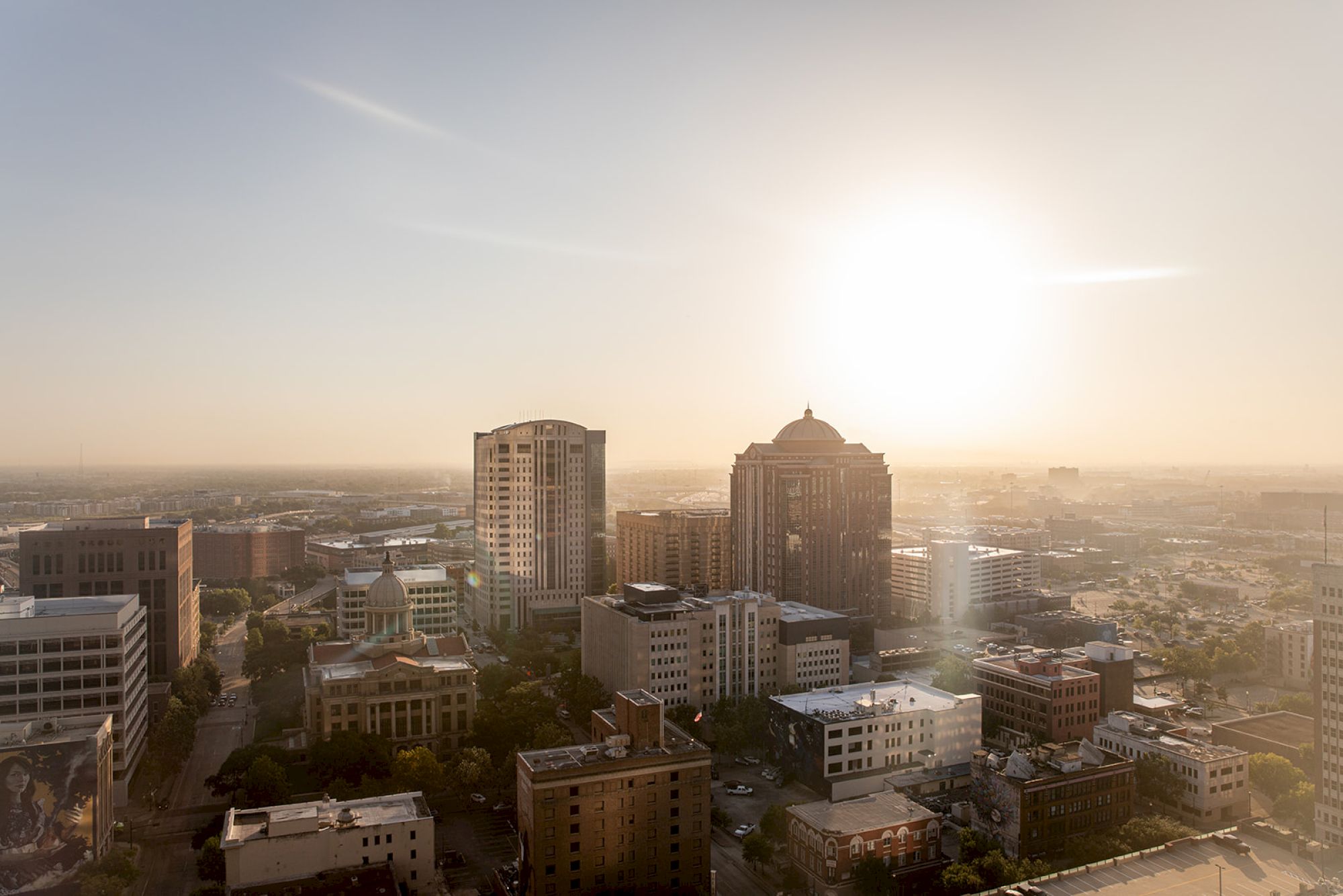A cityscape bathed in warm sunlight with tall buildings under a clear sky. The sun is setting or rising, creating a glowing effect.