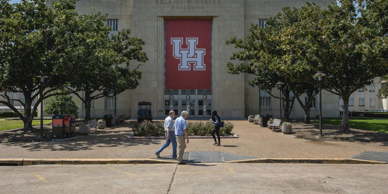 The image shows the front of a building at UH, with trees, people walking, and a large UH logo displayed, under a clear sky.