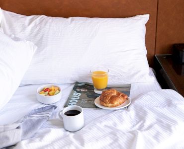 A bed with white sheets, a breakfast tray holding coffee, orange juice, fruit, and a croissant. There's art visible on the wall above.