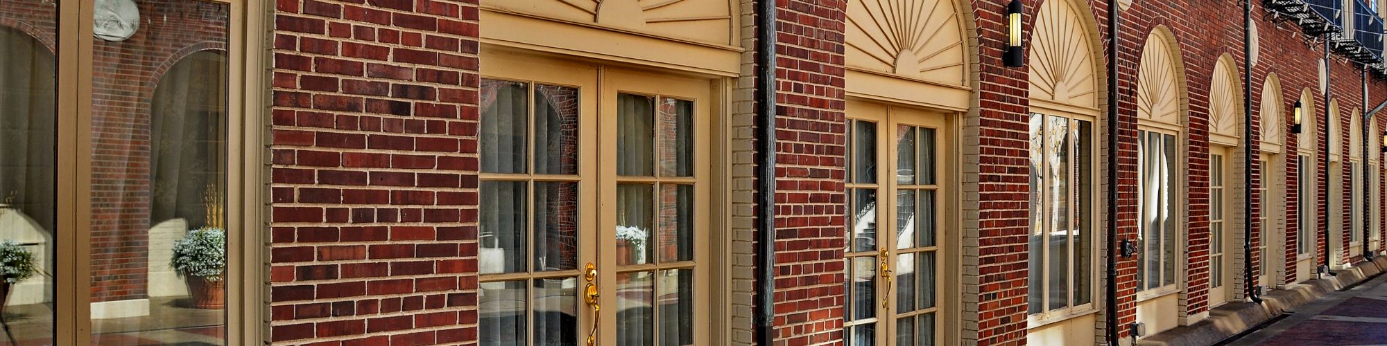 A row of arched windows and doors set in a brick building, featuring balconies above and a paved walkway in front.