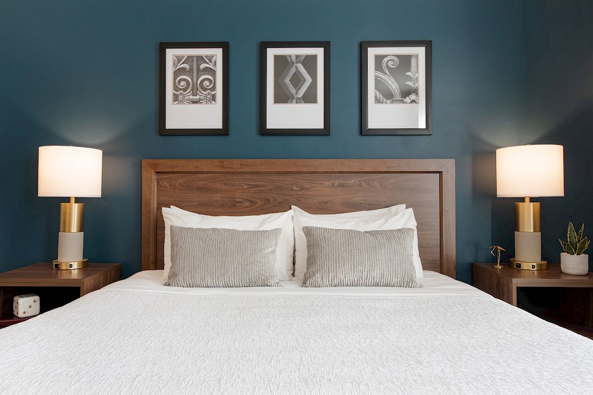 A neatly made bed with two pillows, flanked by lamps on side tables, under three framed pictures on a dark blue wall.