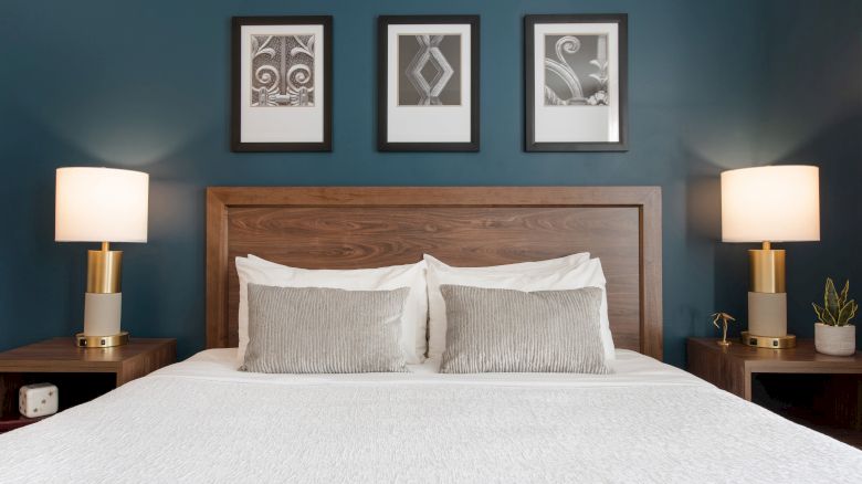 A neatly made bed with two pillows, flanked by lamps on side tables, under three framed pictures on a dark blue wall.