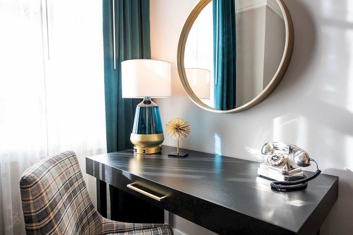 A desk setup with a round mirror, a lamp, a decorative item, an old-fashioned phone, and a plaid chair, in a well-lit room.