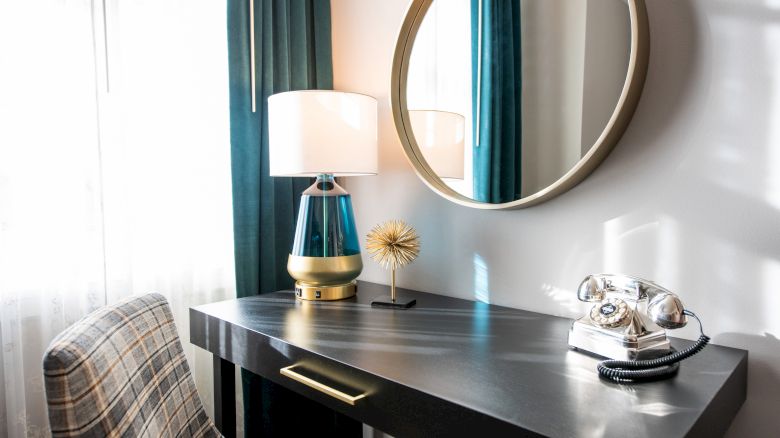 A desk setup with a round mirror, a lamp, a decorative item, an old-fashioned phone, and a plaid chair, in a well-lit room.