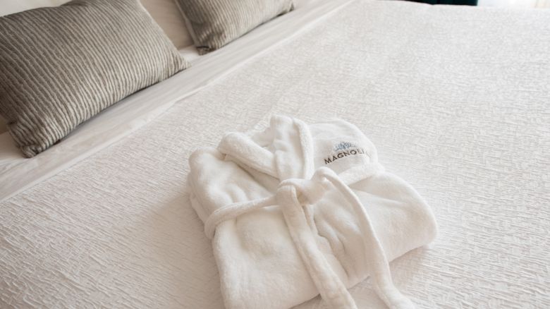A white bathrobe is neatly folded on a made bed with gray pillows in the background.