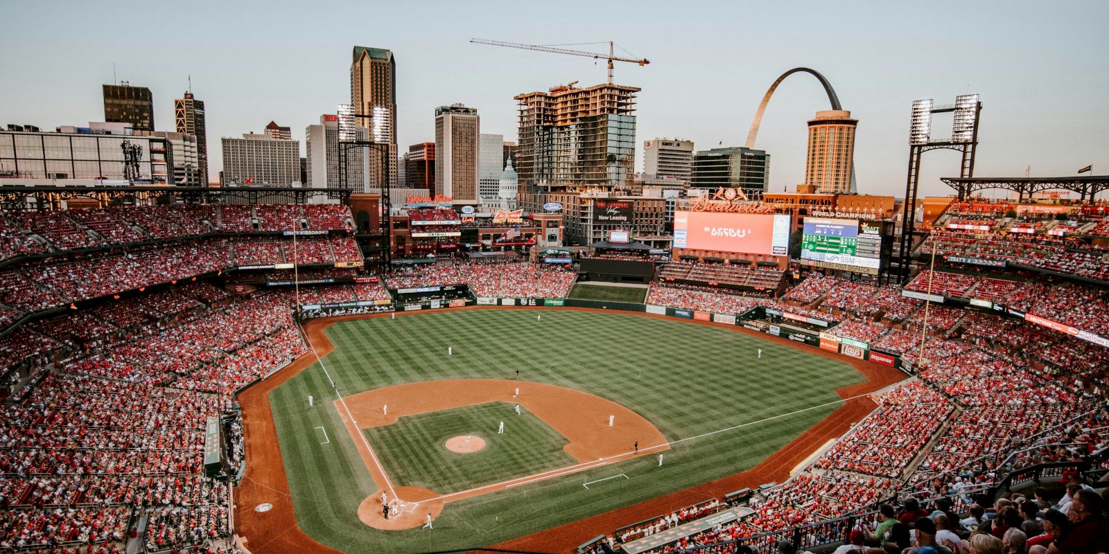 A baseball stadium filled with spectators, a game in progress, with a city skyline and iconic arch in the background on a clear day.