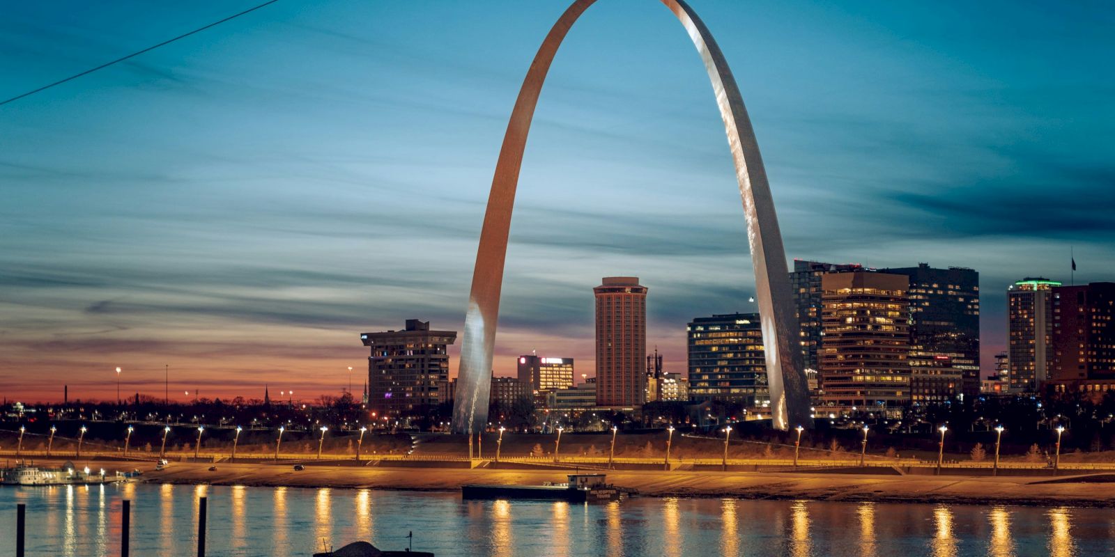 A city skyline at dusk features a large, illuminated arch near a river, under a blue and orange sky.