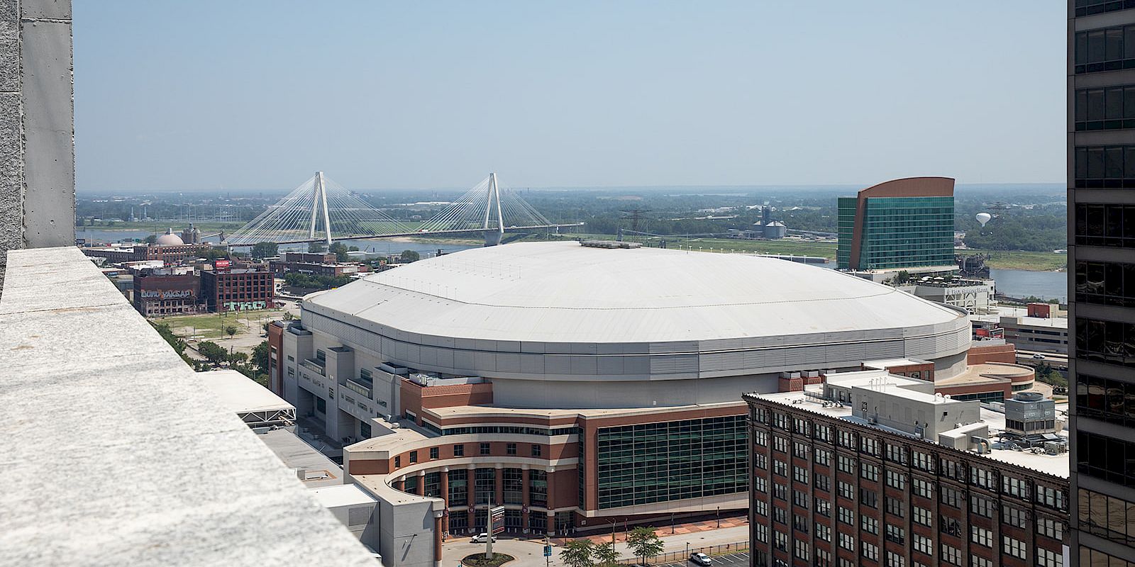 The image shows a large domed stadium surrounded by buildings and a cable-stayed bridge in the background under a clear sky.
