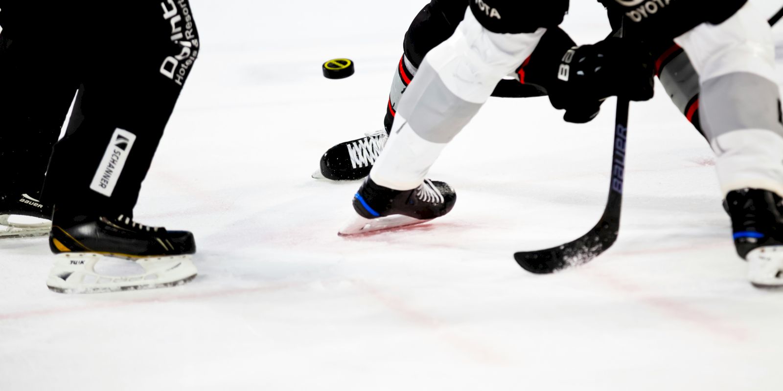 Hockey players on ice, focusing on a puck during a game. Skates and sticks are visible, indicating action in progress.