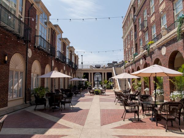 Outdoor courtyard with tables, chairs, and umbrellas, surrounded by brick buildings and overhead string lights under a clear sky.
