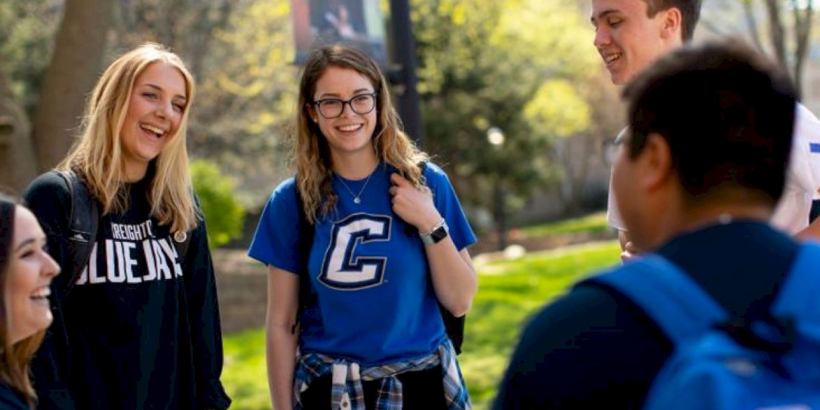 A group of young adults is standing outdoors on a sunny day, wearing casual attire, and engaging in conversation while smiling.