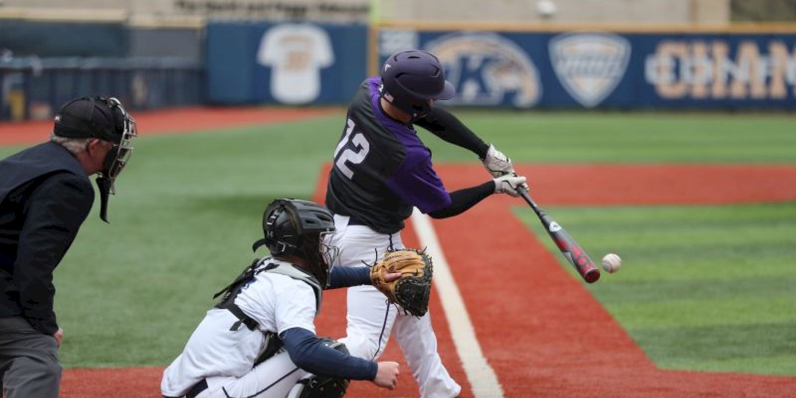 A baseball player in a purple jersey is hitting the ball as a catcher and umpire watch intently at the home plate.