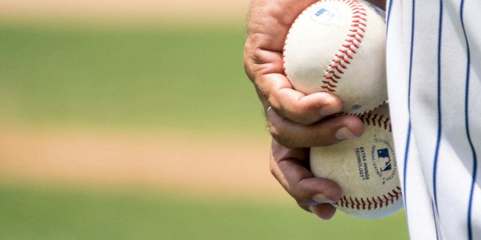 A person in a pinstriped sports uniform holds two baseballs, with a blurry baseball field in the background.