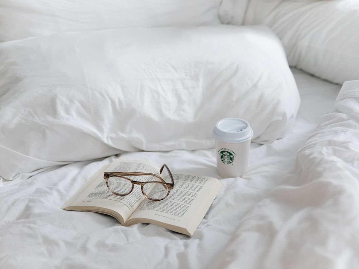 A pair of glasses rests on an open book, beside a Starbucks cup, all on a white bed, creating a cozy, relaxed reading scene.