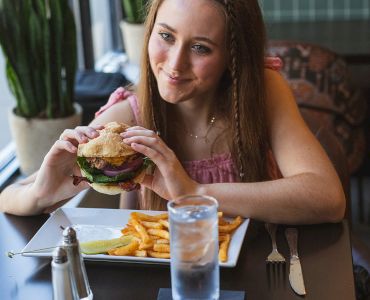 A person is sitting at a table holding a burger. There's a plate of fries and a glass of water nearby. Another person sits across the table.