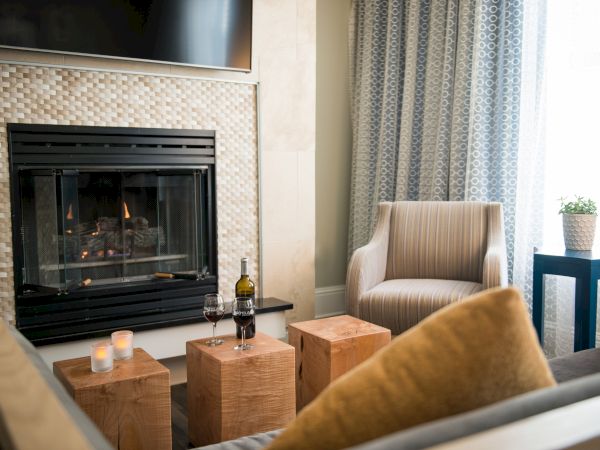 A cozy living room with a fireplace, candles, wooden tables, a bottle of wine, two glasses, and a cushioned chair near a window.