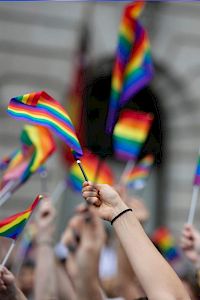 People waving rainbow flags, likely at a pride parade or event, with a blurred building in the background.