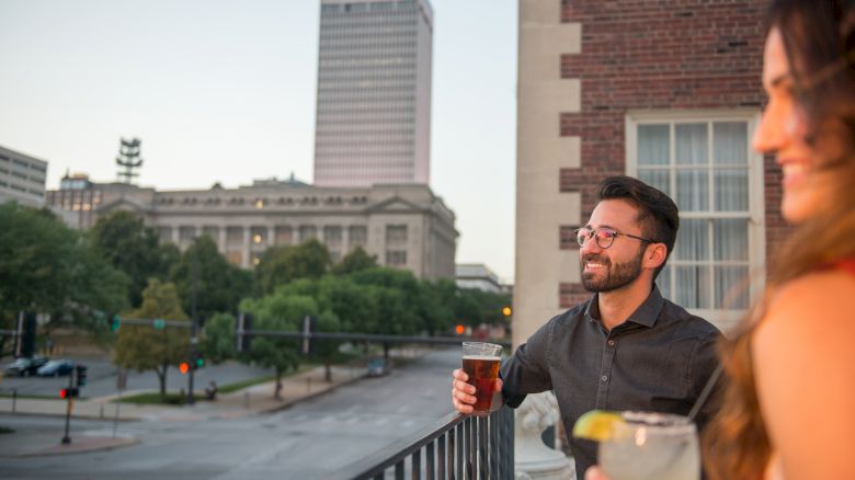 Two people on a balcony enjoy drinks with a cityscape in the background, including a tall building labeled 