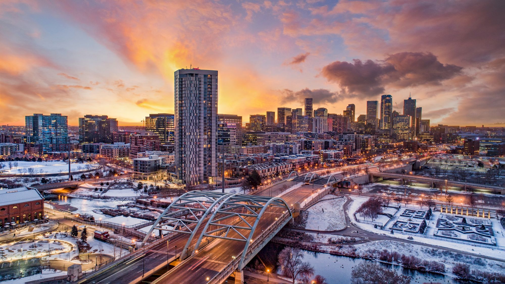 A city skyline at sunset with skyscrapers, a bridge, and snow-covered areas, featuring a vibrant, colorful sky.