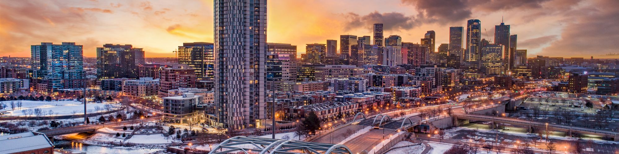 A city skyline at sunset with skyscrapers, a bridge, and snow-covered areas, featuring a vibrant, colorful sky.