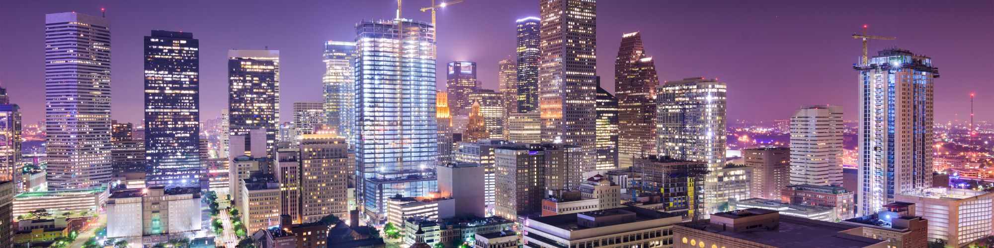 A vibrant city skyline at night with illuminated skyscrapers and construction cranes under a purple sky, surrounded by various buildings.