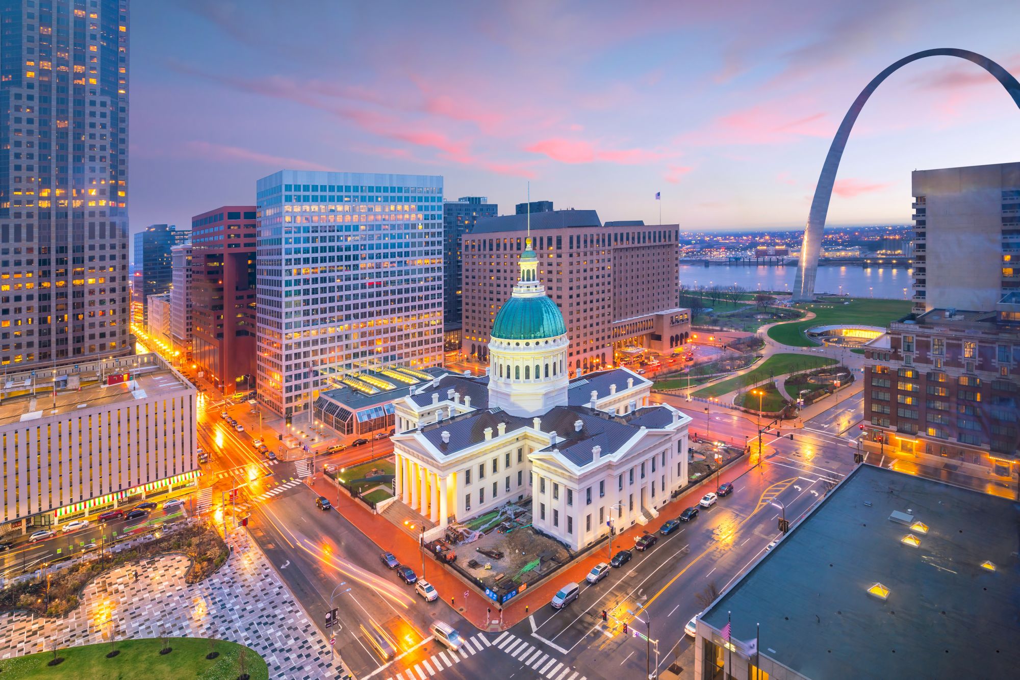 A cityscape featuring the Gateway Arch, historic courthouse, and surrounding buildings at sunset, depicting a vibrant urban area.