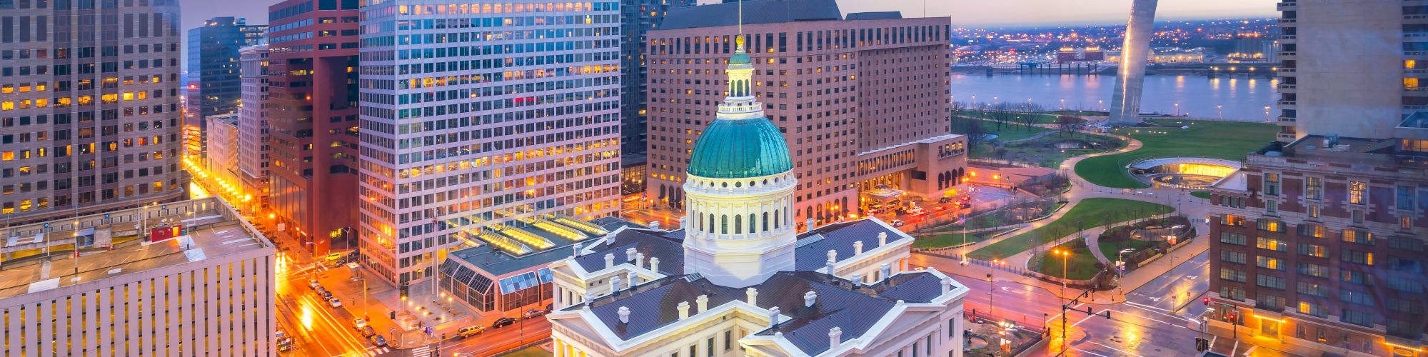 A cityscape featuring the Gateway Arch, historic courthouse, and surrounding buildings at sunset, depicting a vibrant urban area.