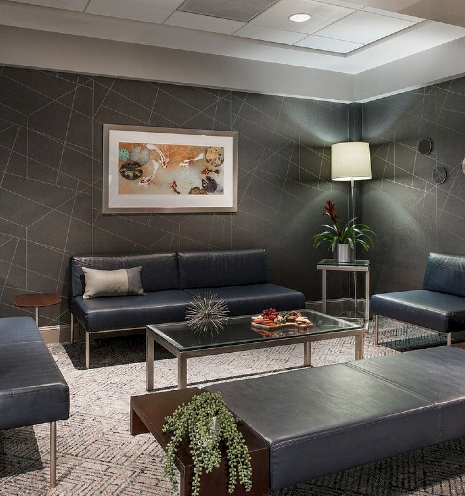 A modern waiting area with dark sofas, plants, art on the wall, and a central glass table with decorative items, warmly lit.