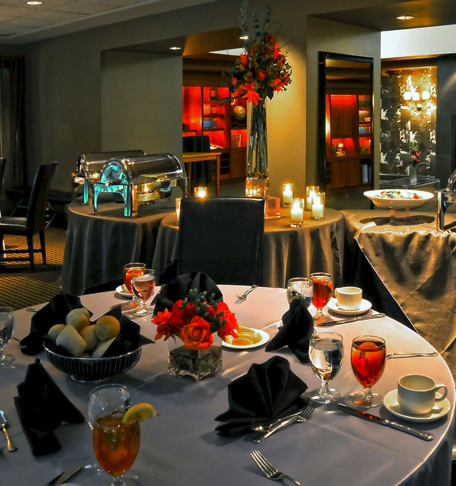 A dining setup with elegantly arranged tables, chafing dishes, candles, and floral centerpieces in a dimly lit room, suggesting an event.