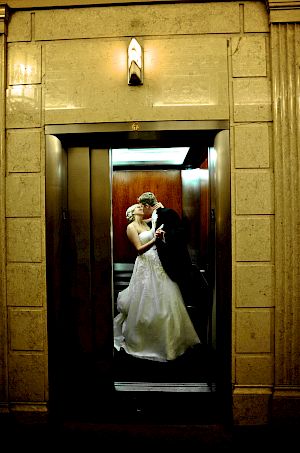 A couple in wedding attire embracing in an elevator with a stone wall and elevator doors.