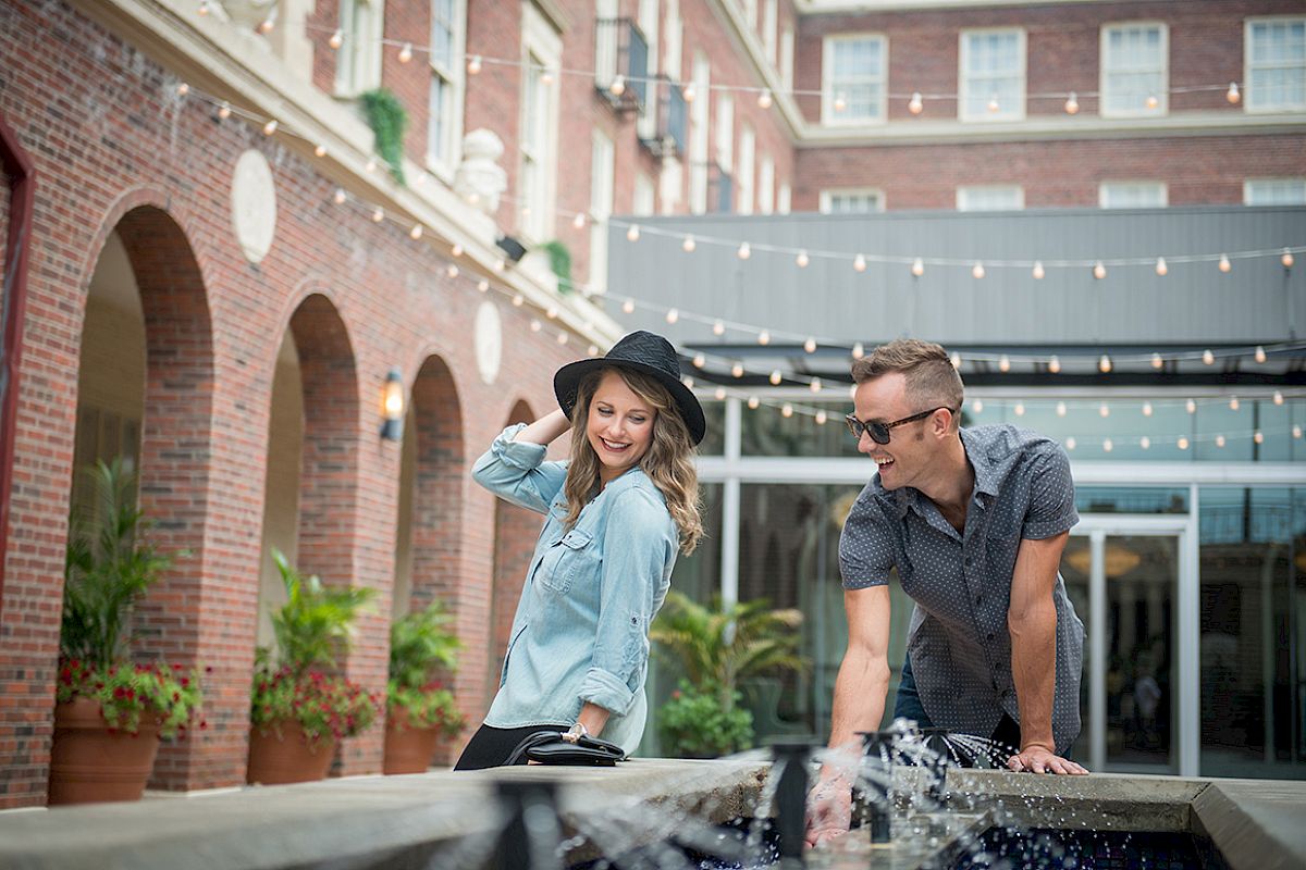 A person in a hat and another in sunglasses near a fountain, enjoying each other's company in a courtyard with string lights above.