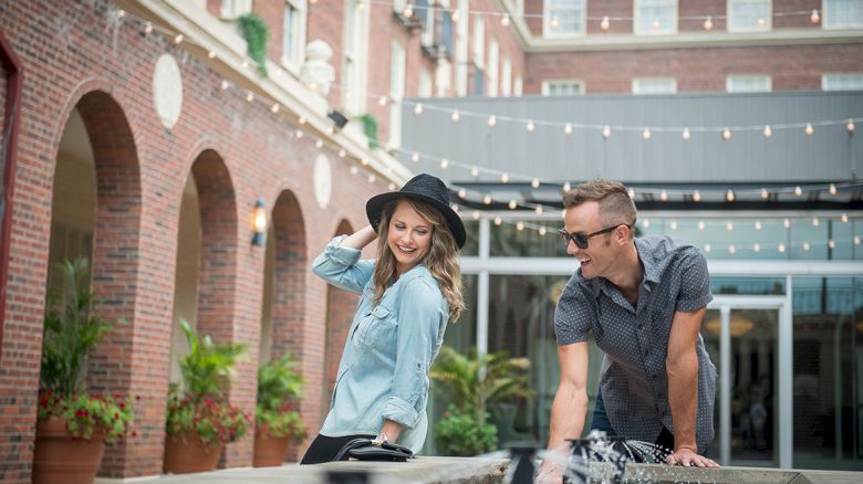 A person in a hat and another in sunglasses near a fountain, enjoying each other's company in a courtyard with string lights above.